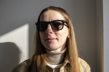 Beyaz bir duvara karşı güneş gözlüklü, ceketli, gülümseyen genç kadın.