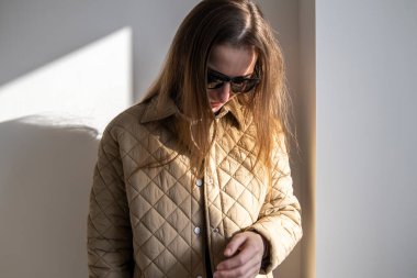 Güneş gözlüklü, üzgün genç bir kadın başını beyaz bir duvarın arkasına eğiyor. 