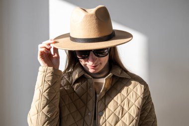 Güneş gözlüklü, şapkasını beyaz bir duvara dayamış genç bir kadın.