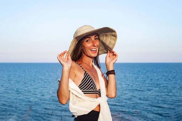 Deniz ve gökyüzünün arka planında şapkalı gülümseyen genç kadın.