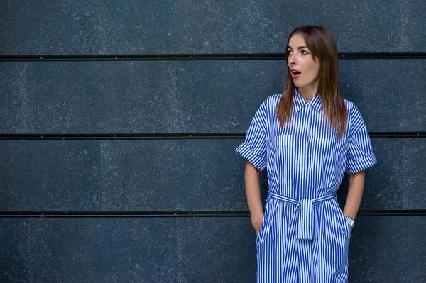 Elbiseli, şaşırmış yüzlü genç bir kadın taş bir duvarın arkasına bakıyor..