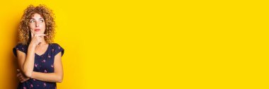 curly young woman pensively looks to the side holding her finger to her chin on a yellow background. Banner