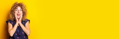 joyful curly young woman in surprise holds her palms on her cheeks on a yellow background. Banner