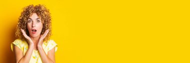 surprised curly young woman holding palms near face on yellow background. Banner.