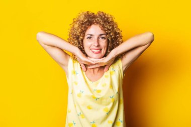 Kıvırcık, gülümseyen genç bir kadın sarı arka planda avuç içlerini çenesinin altında tutuyor..