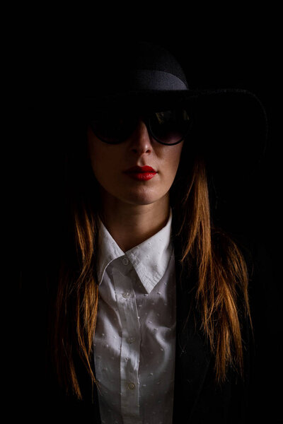 portrait of a beautiful young woman in a black hat and glasses in a dark background.