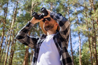 Şapkalı ve gri kareli tişörtlü erkekler, orman arka planındaki dürbünle bakıyorlar..