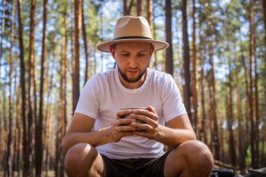 Kamp gezisi sırasında elinde bir kupa sıcak çay tutan şapkalı bir adam. Dağlarda yürüyüş, orman....