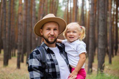 Şapkalı bir adam ormanda yürürken bir çocuğu kollarında tutuyor. Aile dağlara ya da ormana gider..