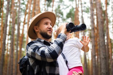 Şapkalı, sırt çantalı bir adam ve bir çocuk ormanda yürürken dürbünle bakıyorlar. Aile dağlara ya da ormana gider..