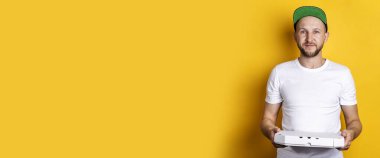 Beyzbol şapkalı gülümseyen genç adam sarı arka planda bir paket pizza tutuyor. Pankart.