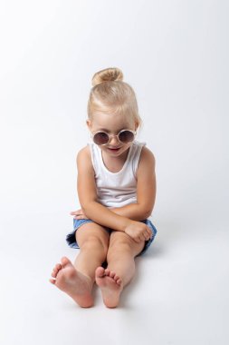 Güneş gözlüklü sarışın çocuk stüdyoda arka planda otururken poz veriyor..