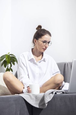 Elinde laptopuyla bir fincan kahve olan genç bir kadın kanepede oturuyor..