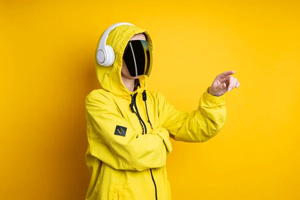 Young woman in cyberpunk glasses with headphones clicks on a virtual screen on a yellow background.