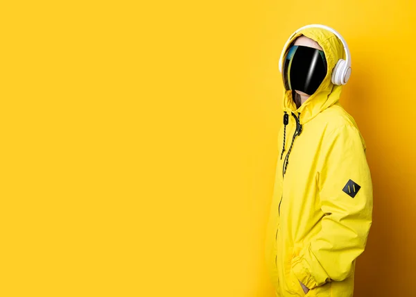 Young woman in cyberpunk glasses mask with headphones in a yellow jacket on a yellow background.