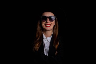 Siyah şapkalı, gözlüklü ve siyah arka planda ceketli gülümseyen bir kadın..