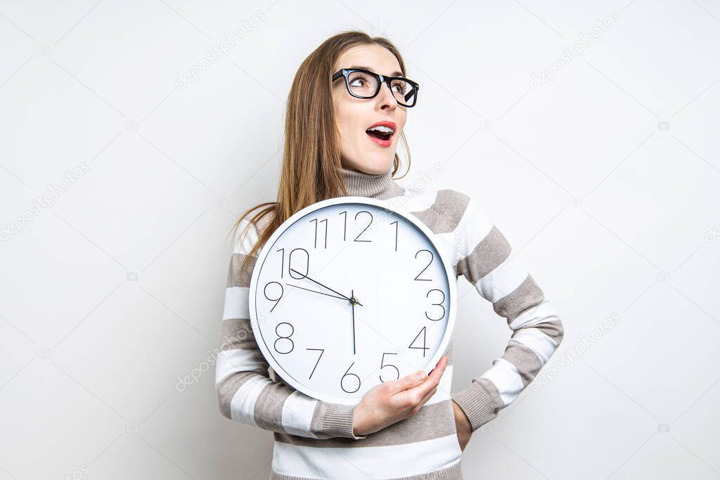 Young woman looks up in surprise holding a clock on a light background 