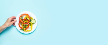 Dişi bir el mavi arka planda doğranmış sebzeli bir tabak tutuyor. Üst manzara, düz yatış. Pankart.