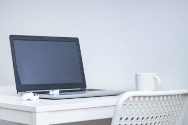 Laptop, fincan, kablosuz kulaklık beyaz bir masada, beyaz sandalye. İşyeri 