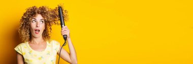 young woman with curly hair straightens her hair in shock looks at a hair straightener on a yellow background. Banner