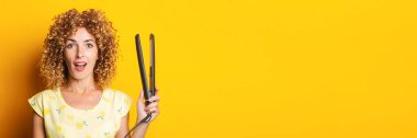 surprised girl with curly hair holding a hair straightener on a yellow background. Banner.
