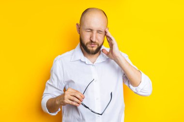 Young man squint taking off his glasses holding his temple on a yellow background.