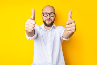 Smiling young man holding thumbs up gesture class on yellow background.