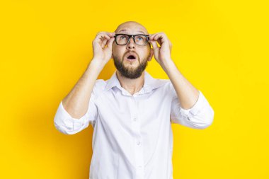 Bald young man holding glasses looks up in surprise on a yellow background.
