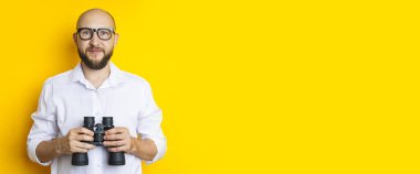 Young man in glasses with binoculars on a yellow background. Banner.