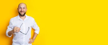 Smiling young man in white shirt holding glasses on yellow background. Banner.
