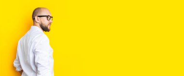 Bald young man with a beard in glasses standing with his back on a yellow background. Banner.