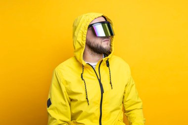 Young man in virtual reality glasses in a yellow jacket looks away on a yellow background.