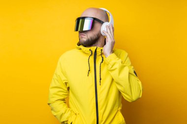 Young man in cyberpunk glasses in a yellow jacket with headphones on a yellow background.