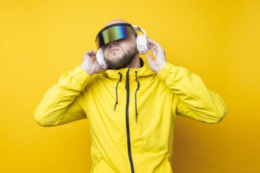 Young man in cyberpunk glasses in a yellow jacket with headphones on a yellow background.