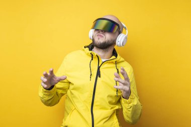 Young man in cyberpunk glasses in a yellow jacket with headphones dancing on a yellow background.