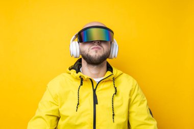 Young man in cyberpunk glasses in a yellow jacket with headphones on a yellow background.