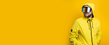 Young man in virtual reality glasses in a yellow jacket looks away on a yellow background. Banner.