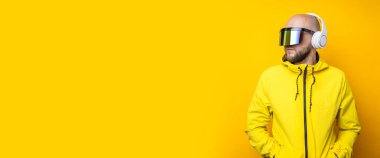 Stylish young man in cyberpunk glasses in a yellow jacket with headphones on a yellow background. Banner.