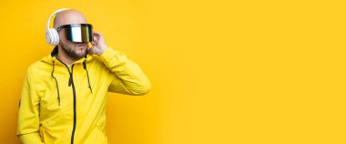 Stylish young man in cyberpunk glasses in a yellow jacket with headphones on a yellow background. Banner.