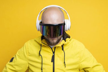 Young man in cyberpunk glasses in a yellow jacket with headphones looks down on a yellow background.