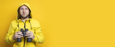 Young man in a yellow jacket holding binoculars on a yellow background. Banner.