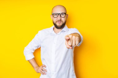 Bald young man in glasses points his finger in front of him on a yellow background.