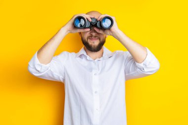 Smiling young man with beard looking through binoculars on yellow background.