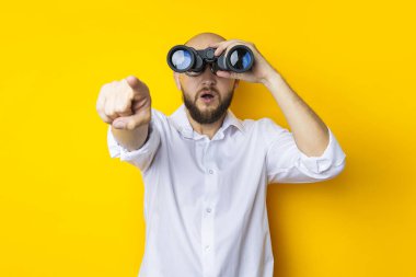 Surprised young man looking through binoculars pointing his finger in front on yellow background.