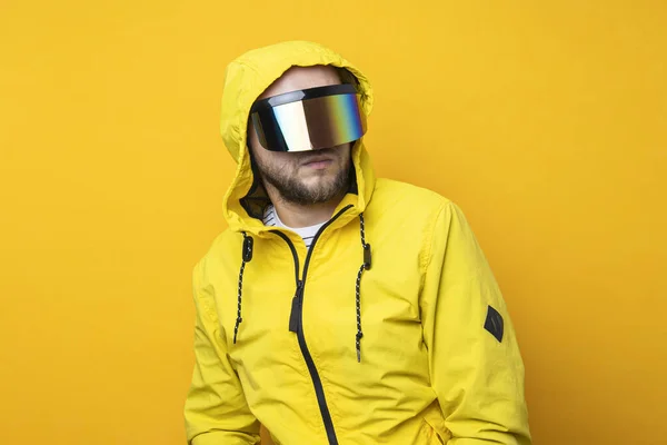 Young man in cyberpunk glasses in a yellow hooded jacket on a yellow background.