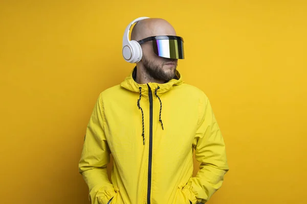 Young man in cyberpunk glasses in a yellow jacket with headphones on a yellow background.