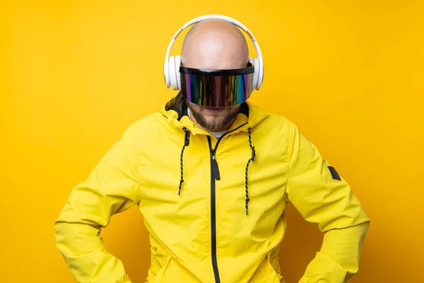Young man in cyberpunk glasses in a yellow jacket with headphones looks down on a yellow background.