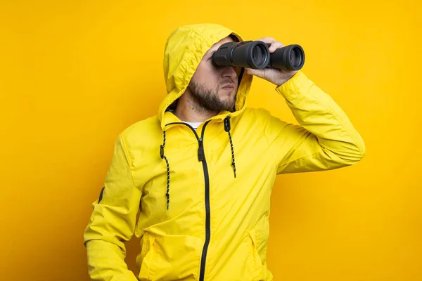 Young man in yellow raincoat looking through binoculars on yellow background.