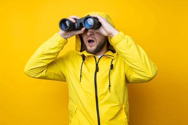 Surprised young man in yellow raincoat looking through binoculars on yellow background.
