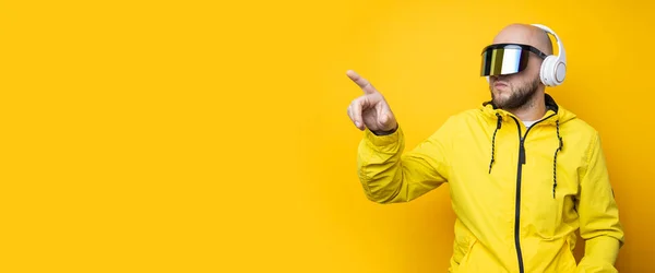 Young man in cyberpunk glasses with headphones clicking on an invisible screen on a yellow background. Banner.